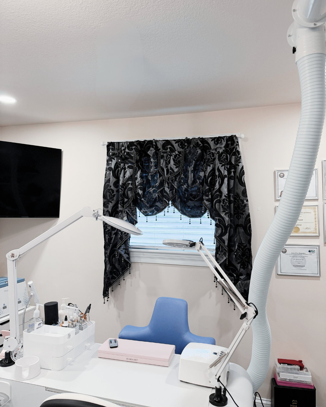 A home-based beauty or nail studio setup featuring a white desk with dual adjustable white arm lamps, a blue ergonomic chair, a pink manicure tray, and a window dressed in black damask curtains.