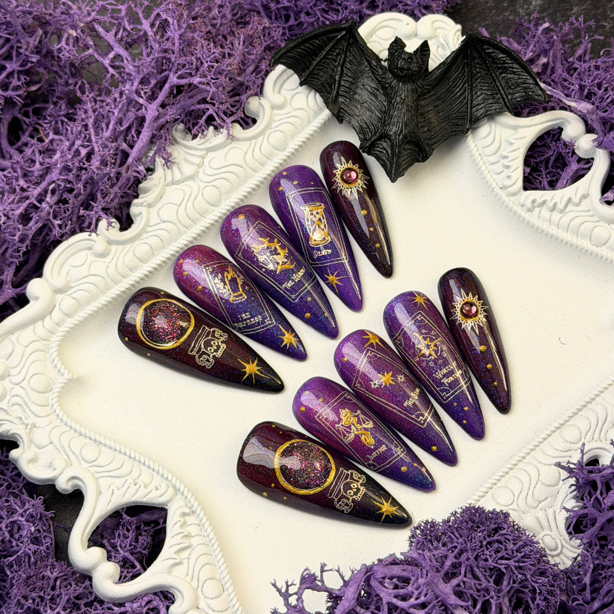 A set of long coffin-shaped nail art press-on nails with purple and black celestial designs, displayed on a white decorative tray with purple moss and a black bat figurine.