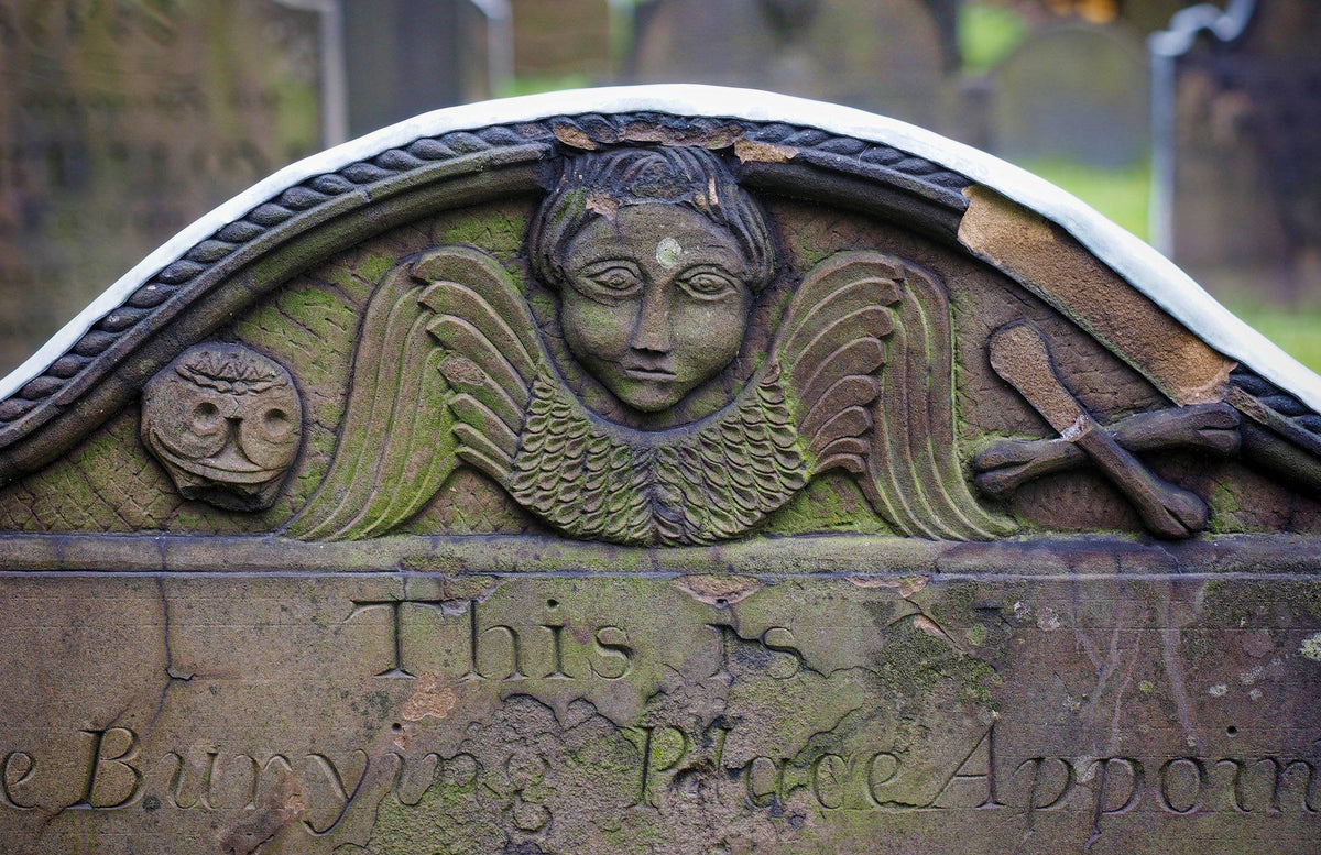 Winged angel carved on a weathered historic gravestone beside a skull and hourglass