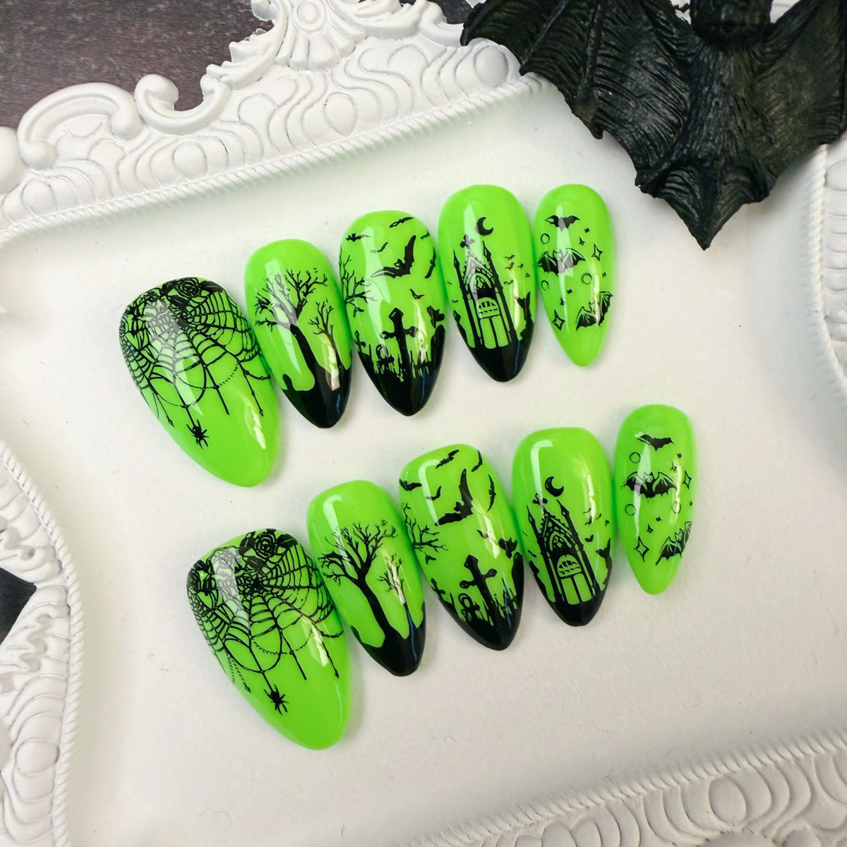 A set of neon green coffin-shaped press-on nails with black Halloween-themed designs including spiderwebs, haunted houses, bats, and tombstones, displayed on a white decorative tray.