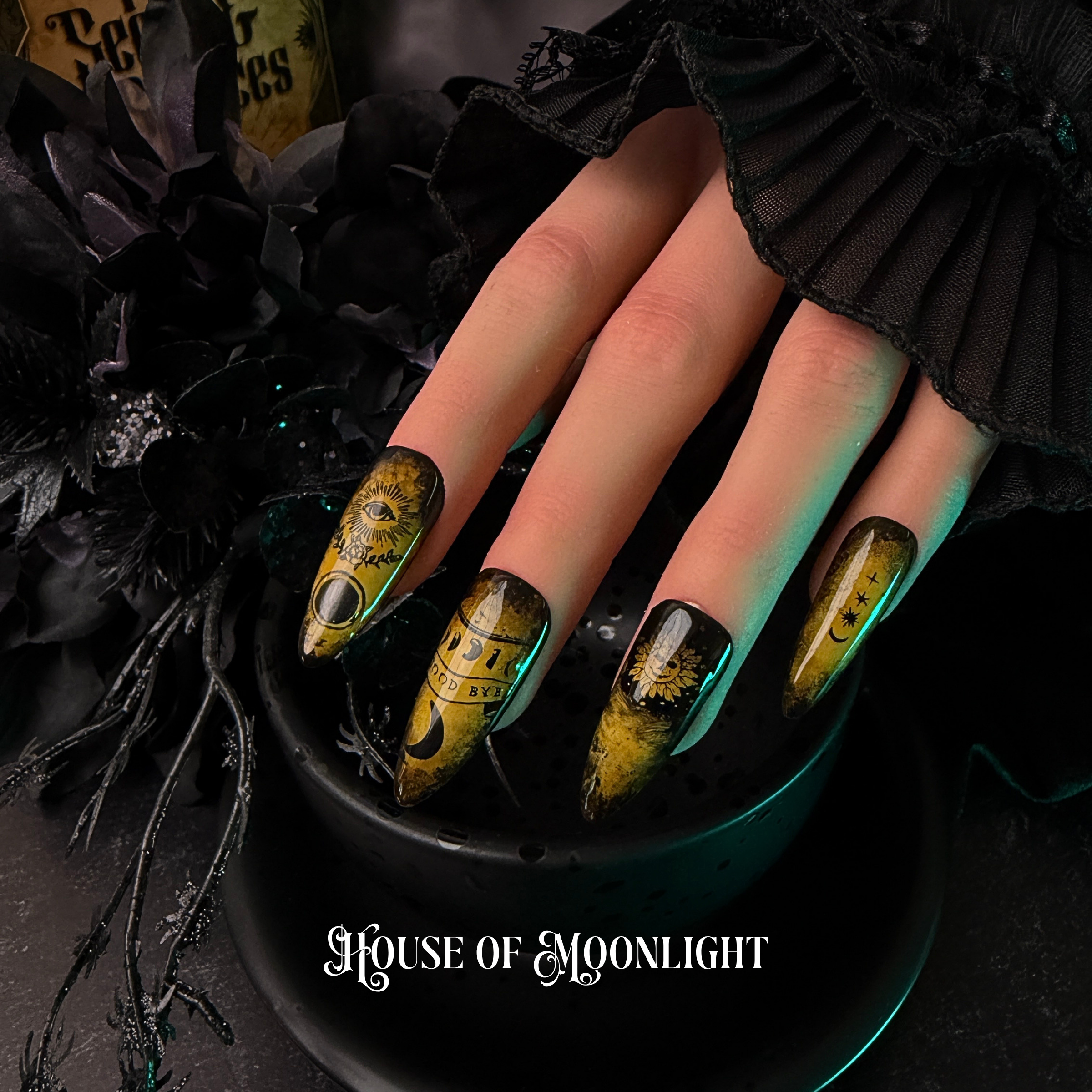 A close-up of a hand with gothic nail art featuring yellow and black celestial ouija designs, resting in a black bowl with the text 'House of Moonlight' at the bottom.
